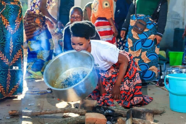 bemba bride in traditional skirt holding pot over a fire while crouched on the ground. Several relatives in colorful attire look on.