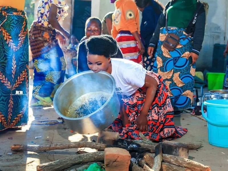 bemba bride in traditional skirt holding pot over a fire while crouched on the ground. Several relatives in colorful attire look on.