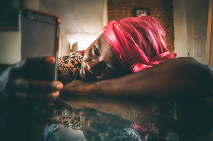 African woman in headwrap leaning against table scrolling on phone, depressed