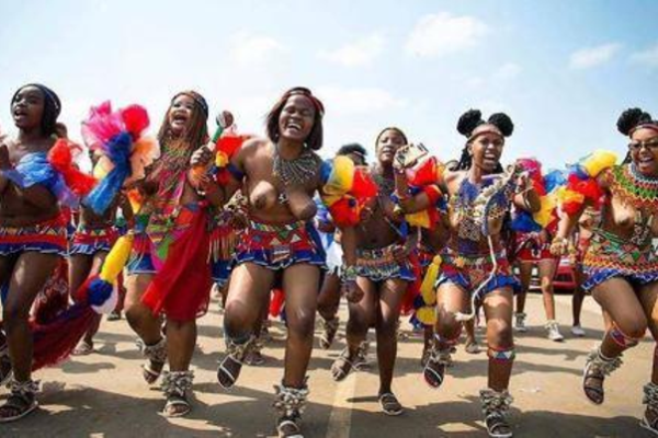 South African women in colorful traditional skirts and bead jewelry, and headdresses. Their sandals are also traditional with embellishments, and they are performing a dance.