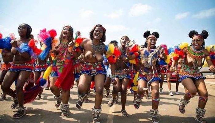 South African women in colorful traditional skirts and bead jewelry, and headdresses. Their sandals are also traditional with embellishments, and they are performing a dance.