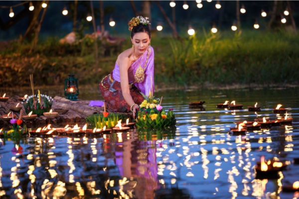 Thai woman in traditional attire and ornate gold and silver foliage headdress placing a basket of flowers in the river. There are floating lights on the water, and fairy lights decorated above.