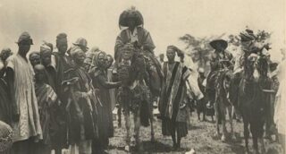 Black and white photo of African people in traditional attire, with a few figures riding horseback. One central figure has a headdress and wearing elaborate robes, with a guard leading him.