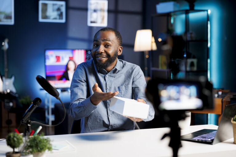 African American man in a home studio with a camera recording himself, presenting a white box