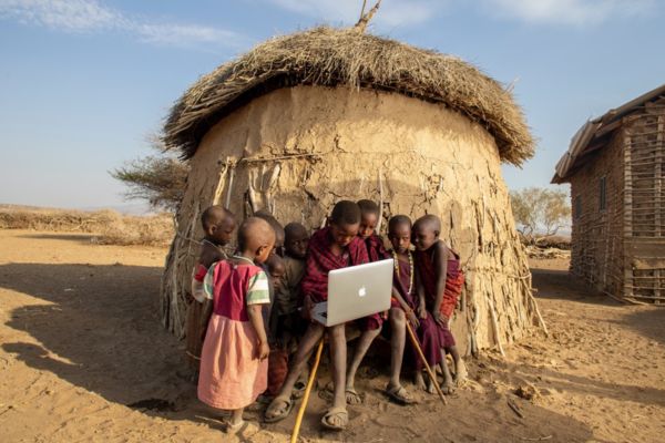 Kenyan children outside of a straw and clay hut gathered around a macbook laptop