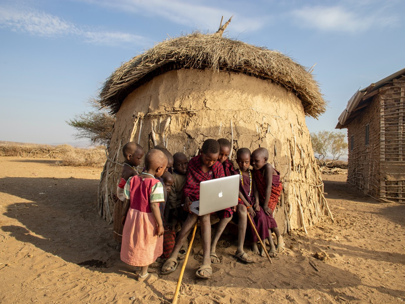 Kenyan children outside of a straw and clay hut gathered around a macbook laptop