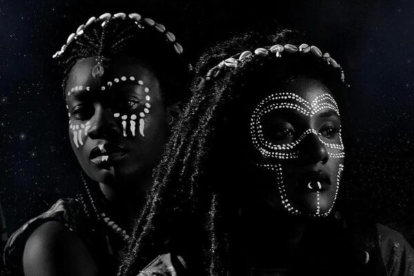 Black and white photograph of African women with hair in dreadlocks decorated with cowry shells, and face paint and other jewelry