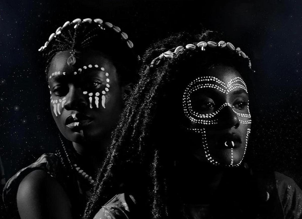 Black and white photograph of African women with hair in dreadlocks decorated with cowry shells, and face paint and other jewelry