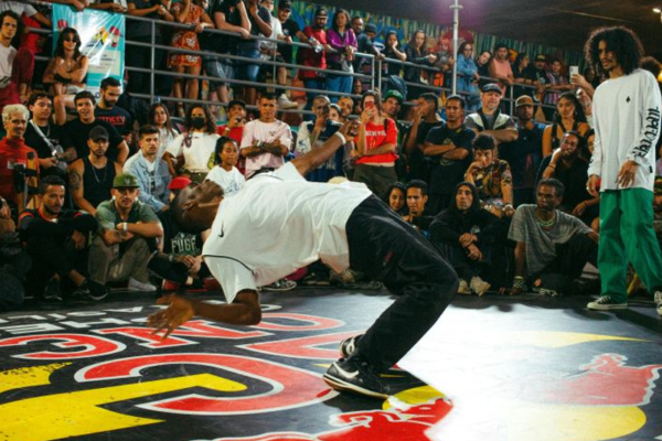 Black break dancer performing in the middle of the floor with onlookers. He is bent backwards and balanced.