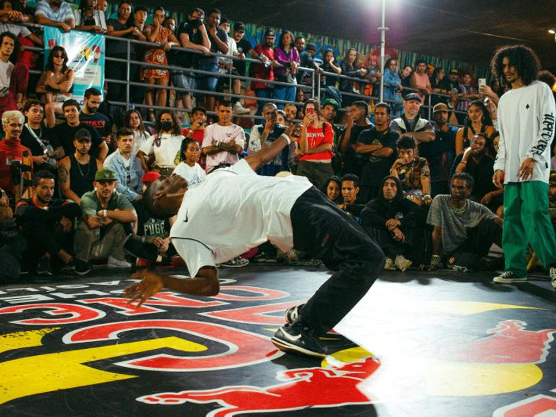 Black break dancer performing in the middle of the floor with onlookers. He is bent backwards and balanced.