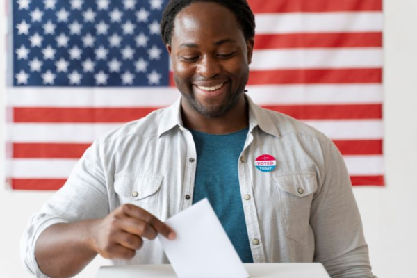 an African American man placing his ballot in the box, with an American flag in the background.