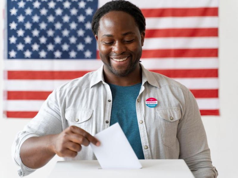 an African American man placing his ballot in the box, with an American flag in the background.