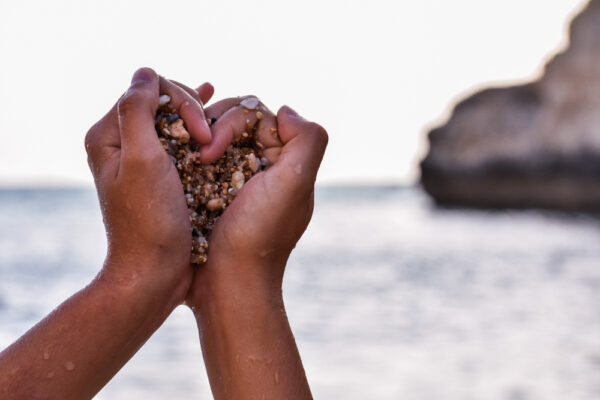 a pair of dark-skinned hands holding pebbles in the shape of a heart in front of the beach