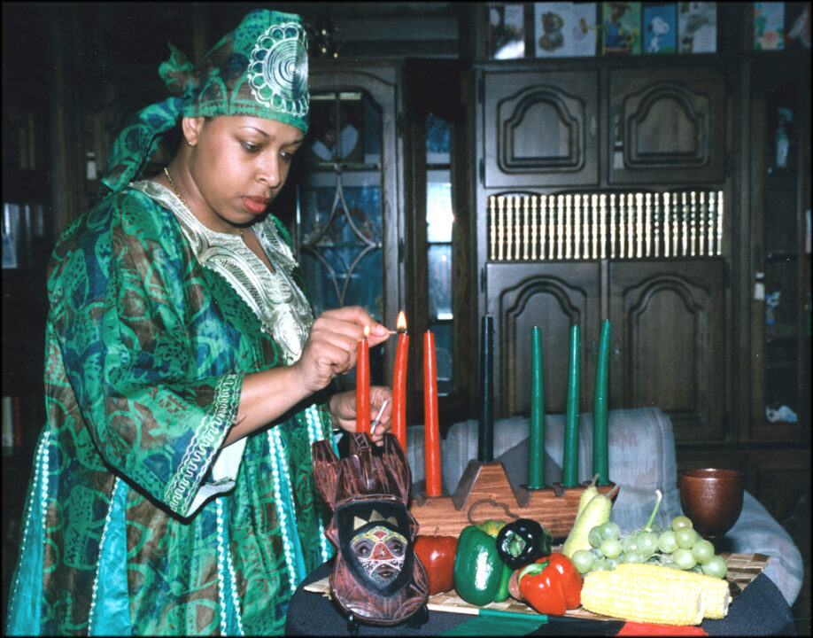 African American woman in traditional attire and matching headscarf lighting the kinara, with the table set up with an African wooden sculpture and fruits