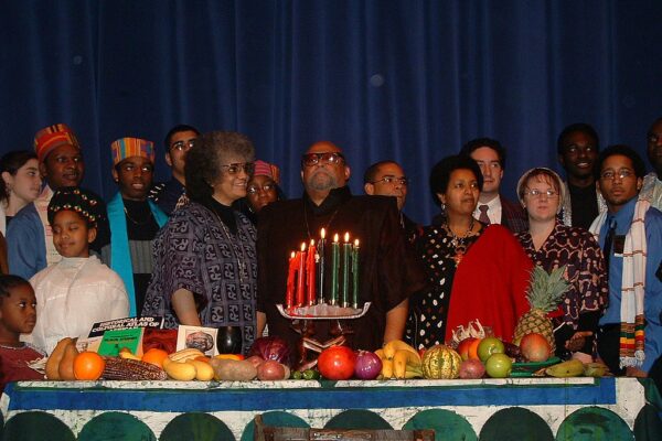 a group of African Americans and a few white people gathered around a table for Kwanzaa, with various fruits spread out with red, green, and black candles lit on a kinara