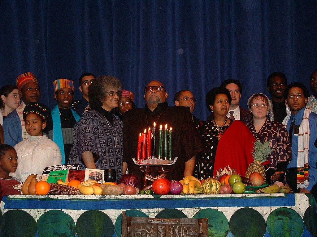 a group of African Americans and a few white people gathered around a table for Kwanzaa, with various fruits spread out with red, green, and black candles lit on a kinara