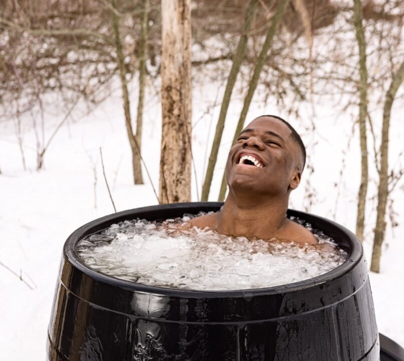 a Black man, smiling, sitting in a barrel of ice water outdoors in the snow.