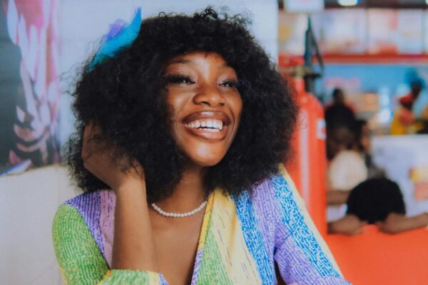 Black woman in colorful dress, smiling ear to ear, sitting in a bright and cheerful cafe