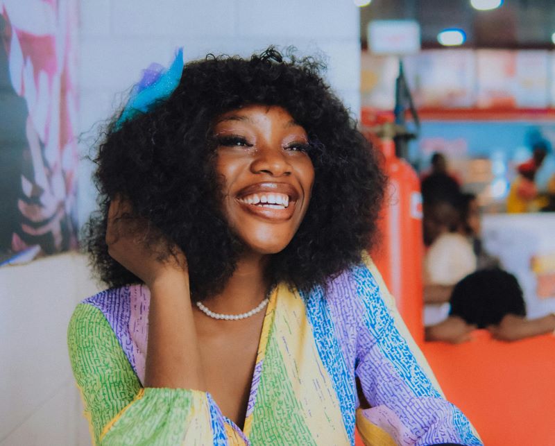 Black woman in colorful dress, smiling ear to ear, sitting in a bright and cheerful cafe