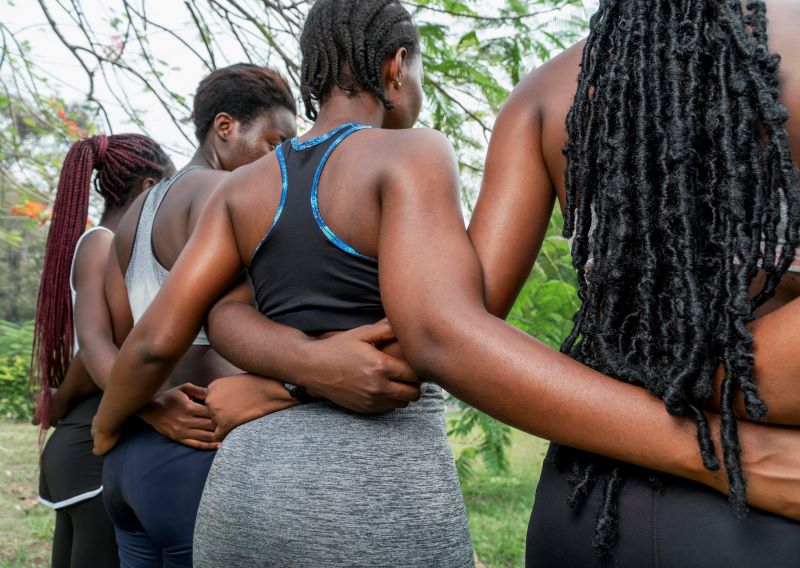 a shot of a group of Black women from behind, with their arms around each other. They are in fitness attire at a park.