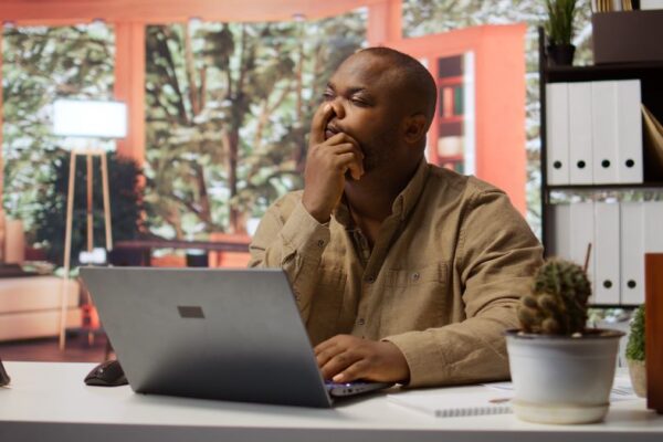 Side Hustles Fueling Black Prosperity In America & Africa In 2025 a Black entrepreneur seated in office contemplating with his finger on his nose while looking at a laptop screen