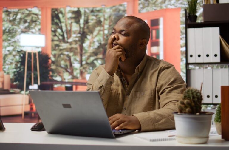 a Black entrepreneur seated in office contemplating with his finger on his nose while looking at a laptop screen