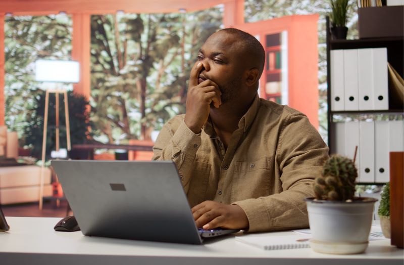 a Black entrepreneur seated in office contemplating with his finger on his nose while looking at a laptop screen