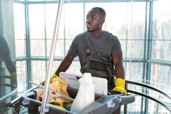a Black man wearing rubber gloves and overalls pushing a cart full of cleaning tools in an office building