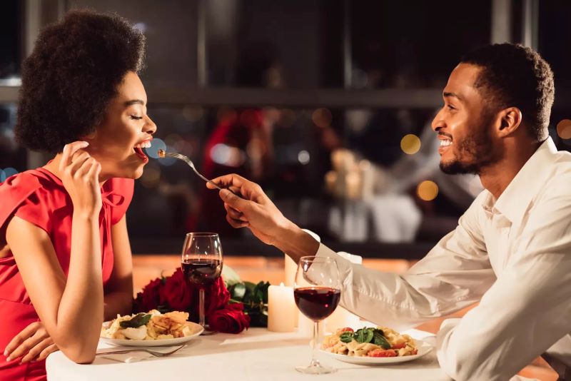African American couple enjoying a candlelit dinner, with the man bringing a fork to the woman's mouth.