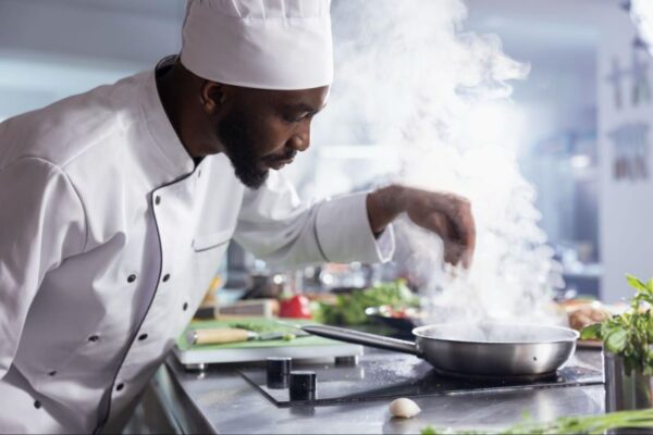 African American chef sprinkling seasoning into a frying pan on a stovetop.