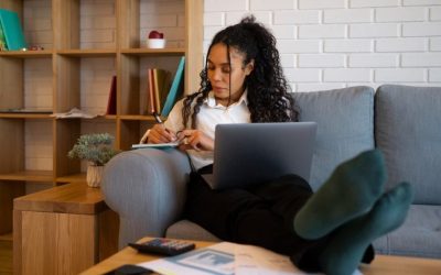 Black woman sitting on couch and writing in a notebook with laptop open