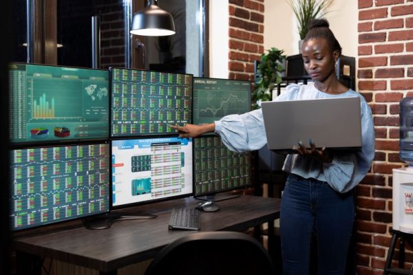woman pointing to stock charts on six screens while holding a laptop