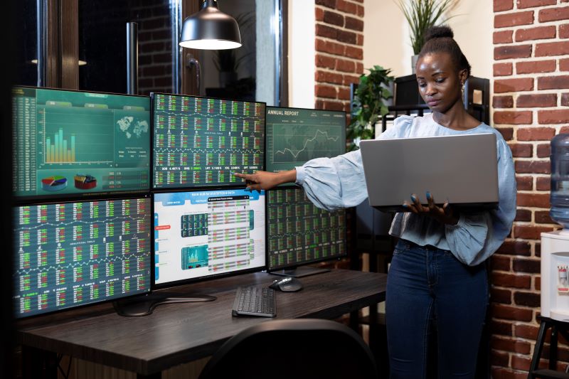 woman pointing to stock charts on six screens while holding a laptop