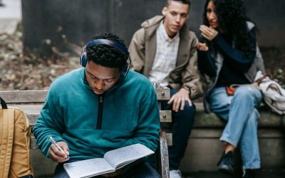 Black student wearing headphones and studying at a bench outdoors while two other students sit behind him and gossip