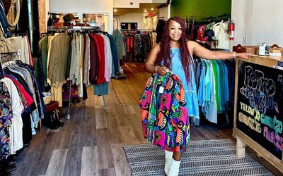 African American woman in a dress and boots holding up clothing in a boutique