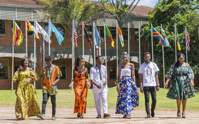 African students on a college campus wearing various traditional patterned formal dress. There are flags of various nations raised behind them in front of a campus building.
