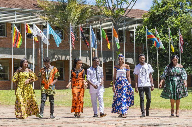African students on a college campus wearing various traditional patterned formal dress. There are flags of various nations raised behind them in front of a campus building.